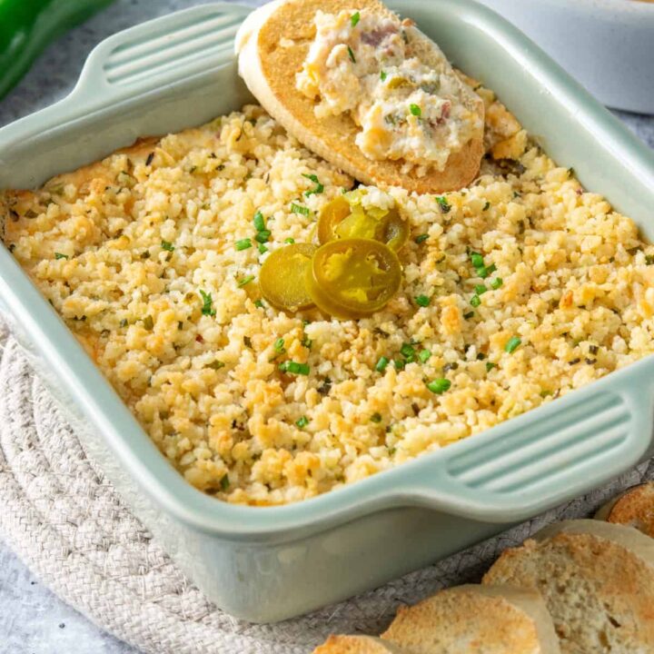 Jalapeno Popper Dip in a baking dish, garnished with jalapeno pepper slices. Theres a fresh jalapeno pepper, toasted french bread slices and crackers next to the dip.