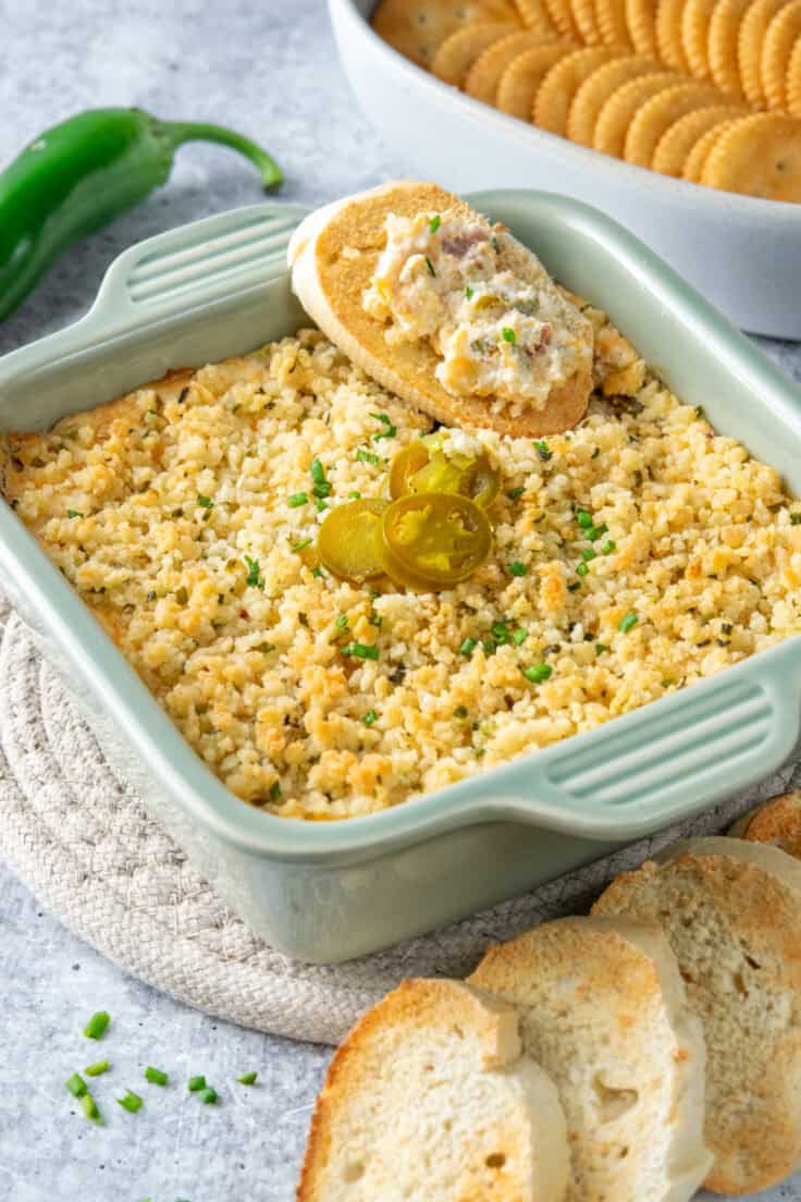 Jalapeno Popper Dip in a baking dish, garnished with jalapeno pepper slices. Theres a fresh jalapeno pepper, toasted french bread slices and crackers next to the dip.