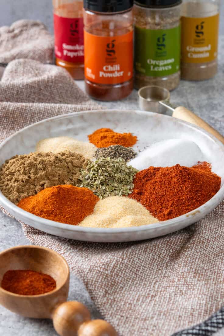 Measured spices for homemade fajita seasoning arranged on a plate with spice bottles in the background.