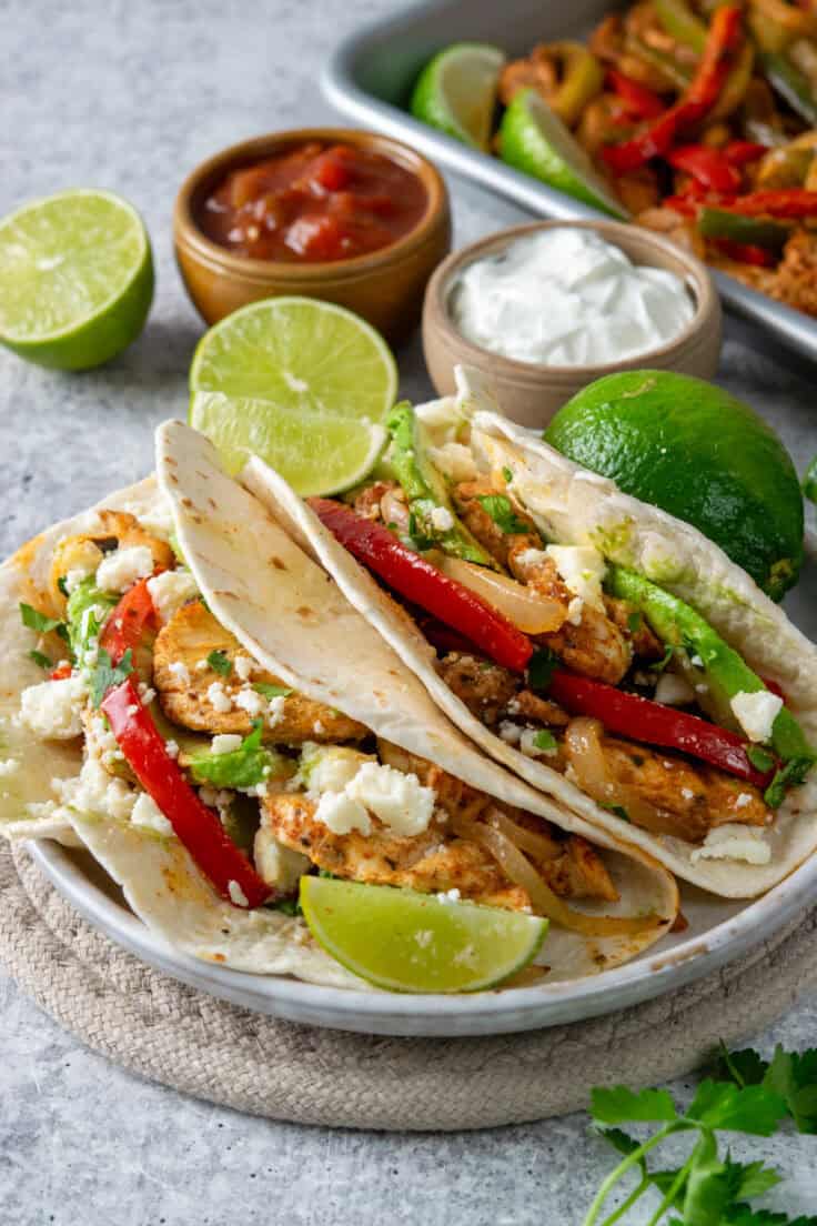 Two chicken fajitas topped with peppers, onions, avocado, fresco cheese, cilantro, and lime next to a sheet pan with extra fajita filling and bowls of salsa and sour cream.