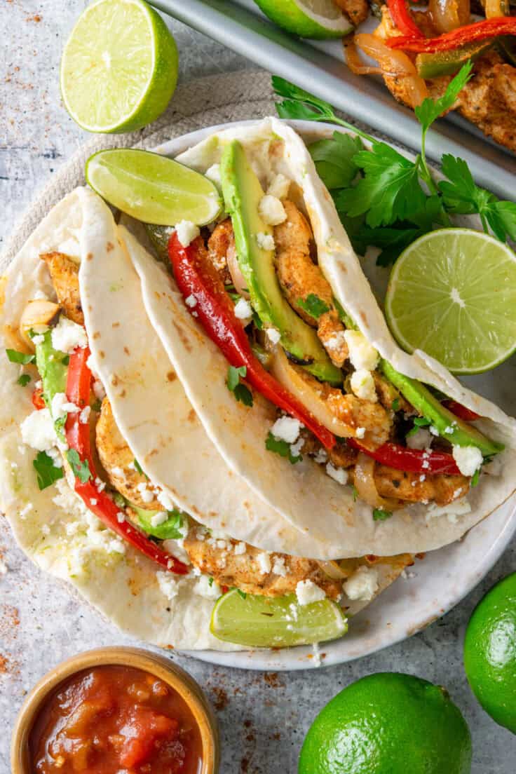 Chicken fajitas in flour tortillas with caramelized peppers and onions, avocado, fresco cheese, and cilantro, with cooked fajita filling on a sheet pan in the background.