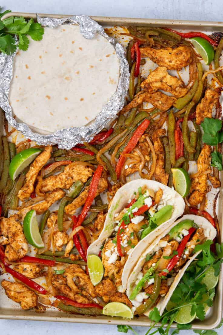 Sheet pan chicken fajitas just out of the oven with roasted chicken, peppers, and onions, foil-wrapped tortillas, and assembled fajitas with toppings and lime.