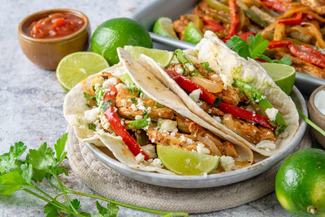 Two chicken fajitas loaded with toppings on a plate next to a sheet pan of cooked chicken, peppers, and onions with bowls of salsa and sour cream.