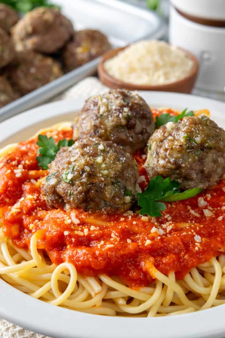 Three baked meatballs on top of spaghetti with marinara sauce, which is next to a tray of the homemade ground beef meatballs.