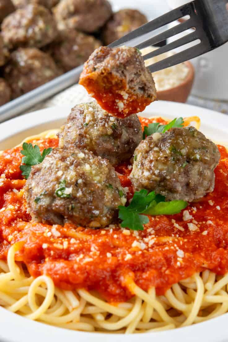Fork lifting half a meatball covered in sauce from a plate of spaghetti and meatballs, with a tray of baked meatballs and parmesan cheese in the background.