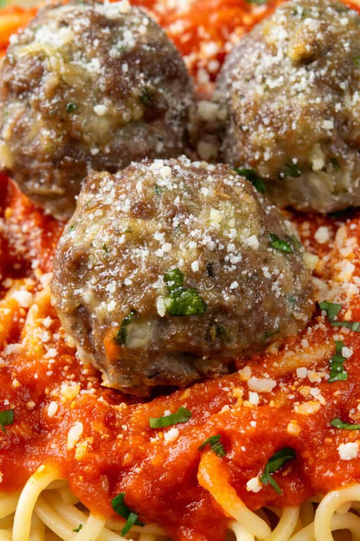 A close-up of the most delicious moist, baked meatballs filled with garlic and herbs with extra grated parmesan on top. The meatballs are on top of a saucy spaghetti.