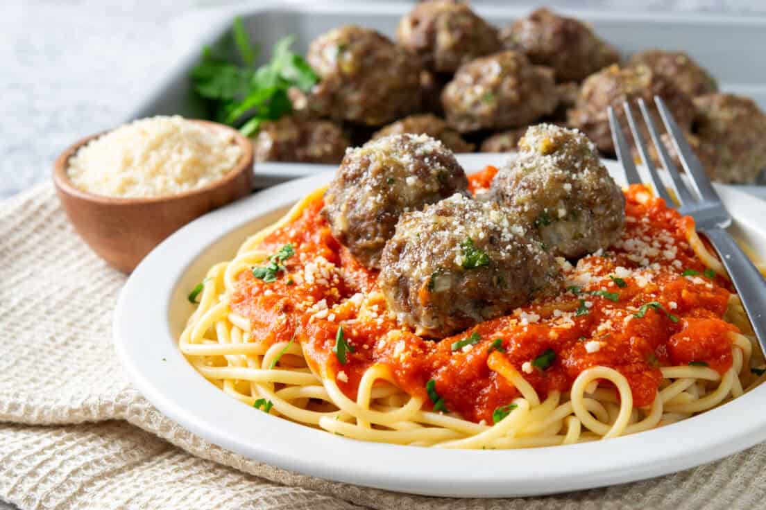 Baked meatballs on a plate piled high with spaghetti and sauce. Next to the spaghetti and meatballs is a sheet pan with more baked meatballs and a small bowl with grated parmesan.