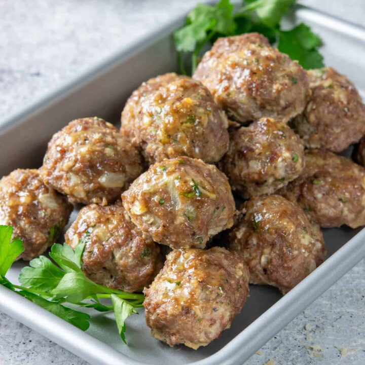 Baked meatballs on a sheet tray with a garlic bulb and small bowl of grated parmesan cheese in the background.