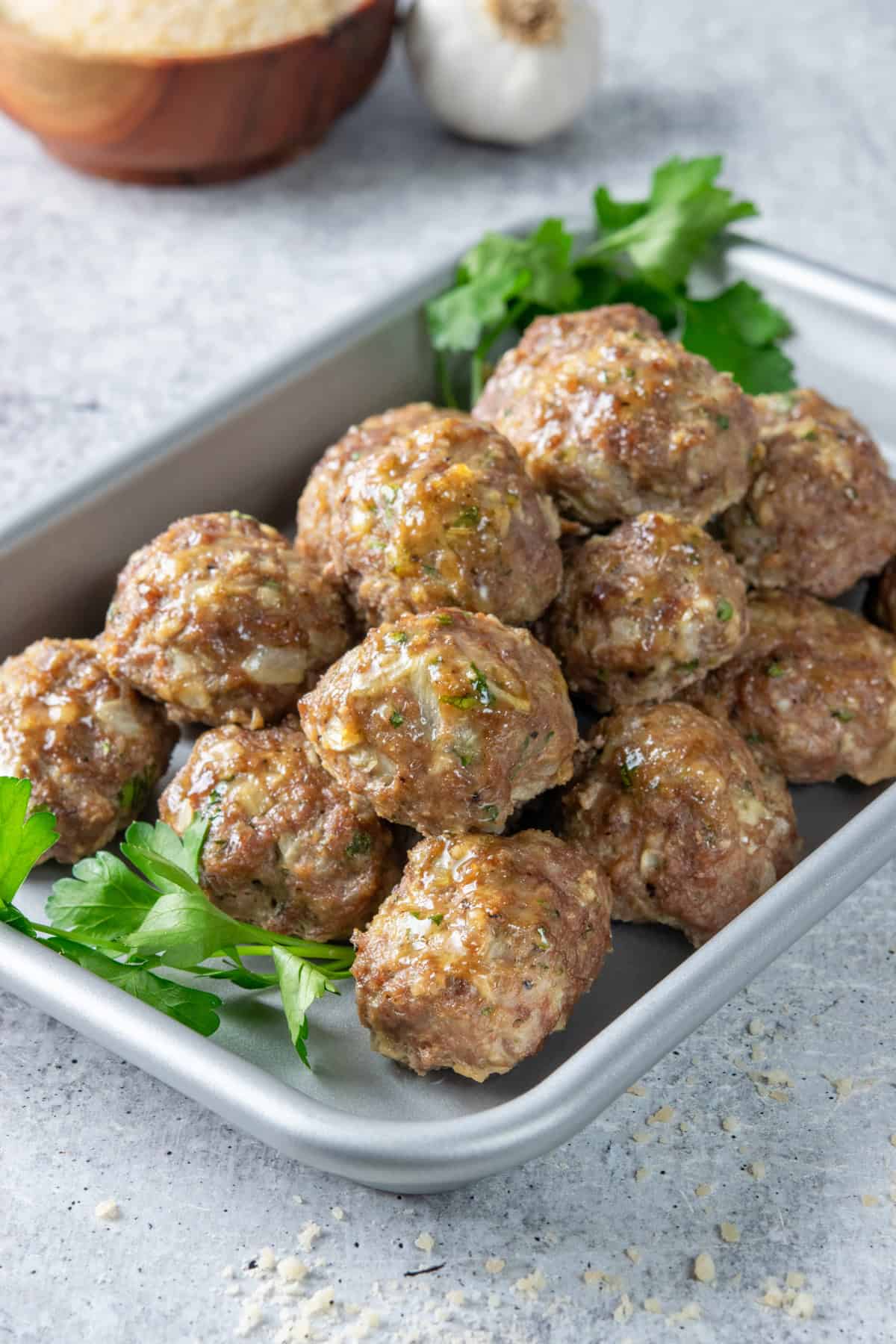 Baked meatballs on a sheet tray with a garlic bulb and small bowl of grated parmesan cheese in the background.