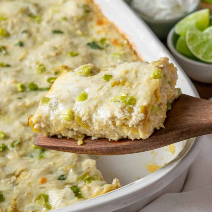 Green chile chicken enchilada casserole slice being lifted from baking dish showing layers of corn tortillas, shredded chicken, green chiles and melted cheese