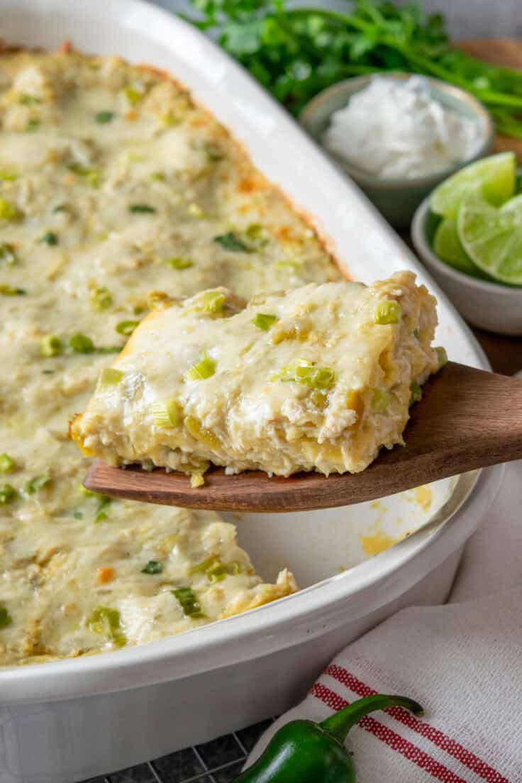 Green chile chicken enchilada casserole slice being lifted from baking dish showing layers of corn tortillas, shredded chicken, green chiles and melted cheese