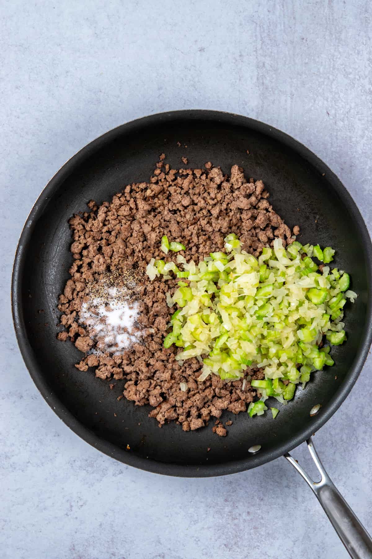 Sautéed onions and celery stirred into browned ground beef with salt and pepper in a skillet.