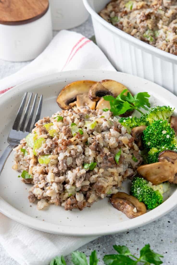 A white plate featuring a serving of hamburger rice casserole paired with vibrant steamed broccoli and savory sautéed mushrooms, highlighting a complete 30-minute ground beef dinner.