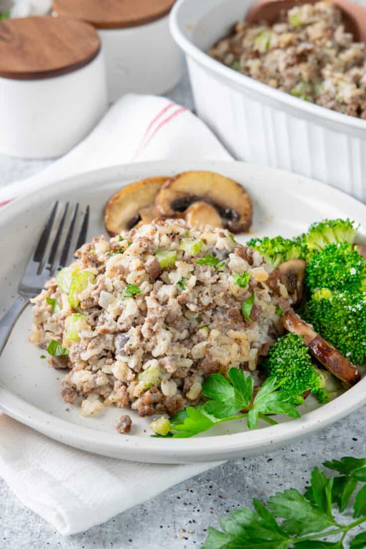 A serving of old-fashioned hamburger rice casserole with a side of broccoli on a plate, positioned next to the full white ceramic casserole dish to show the 4-serving portion size.
