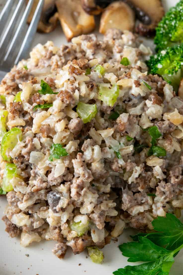 A close-up of a creamy hamburger and rice hotdish serving. The texture shows caramelized ground beef, fluffy rice, and tiny flecks of green celery in a rich cream of mushroom sauce.