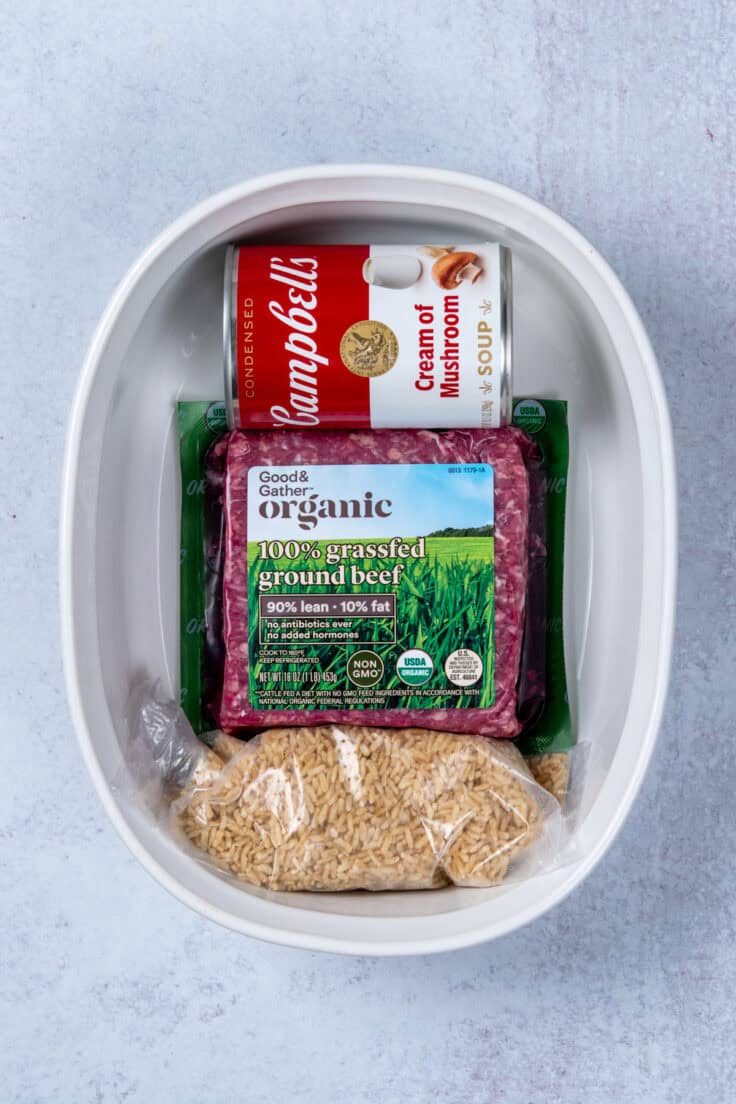 Overhead view of the key ingredients for an easy hamburger rice casserole: a bag of uncooked rice, a package of raw ground beef, and a can of cream of mushroom soup next to a white ceramic baking dish.