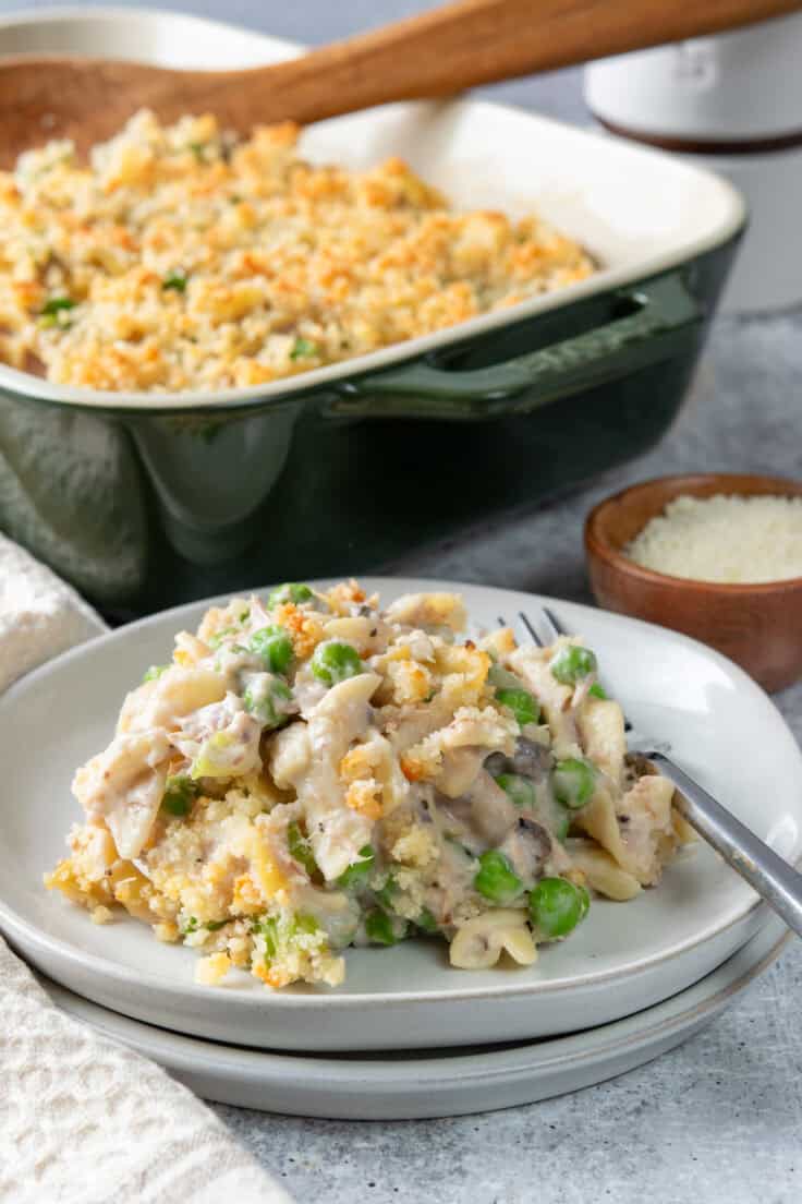 A serving of tuna casserole with a golden parmesan topping next to the casserole dish.
