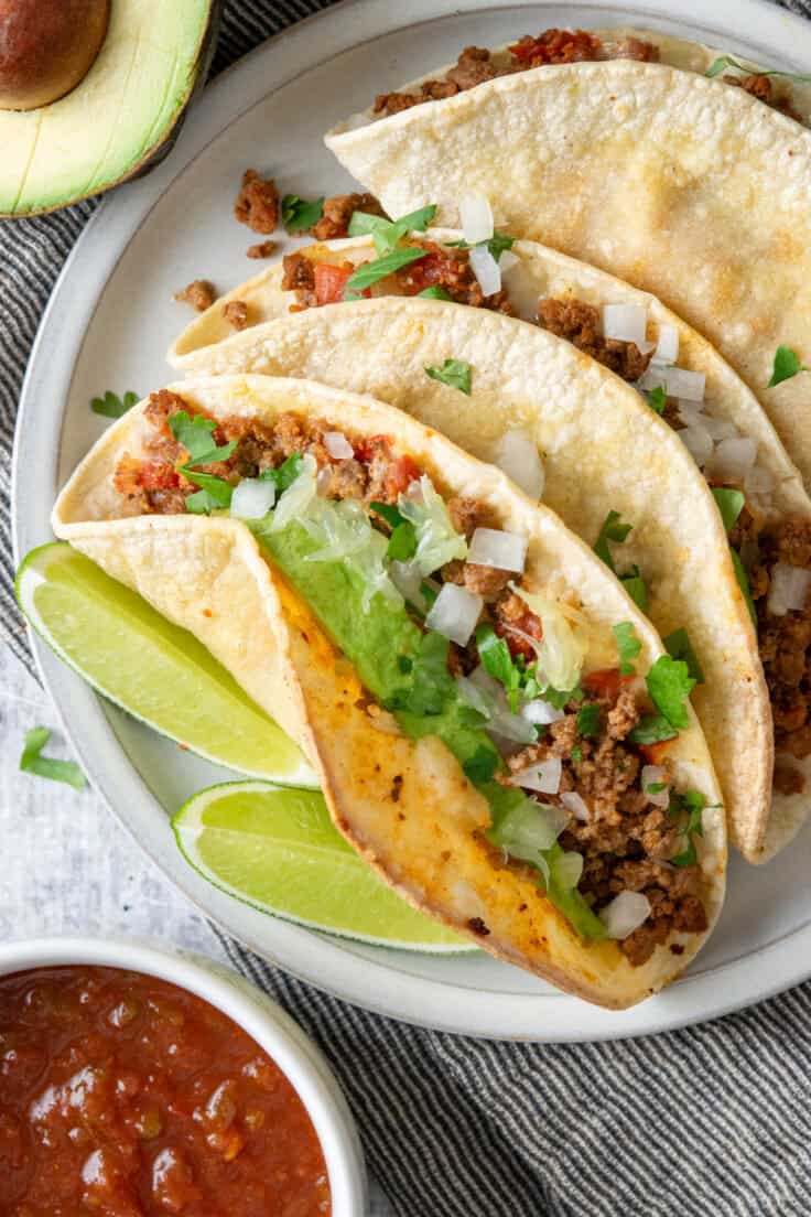 Overhead view of baked ground beef tacos with melted cheese, onion, cilantro, and avocado served with salsa and avocado.