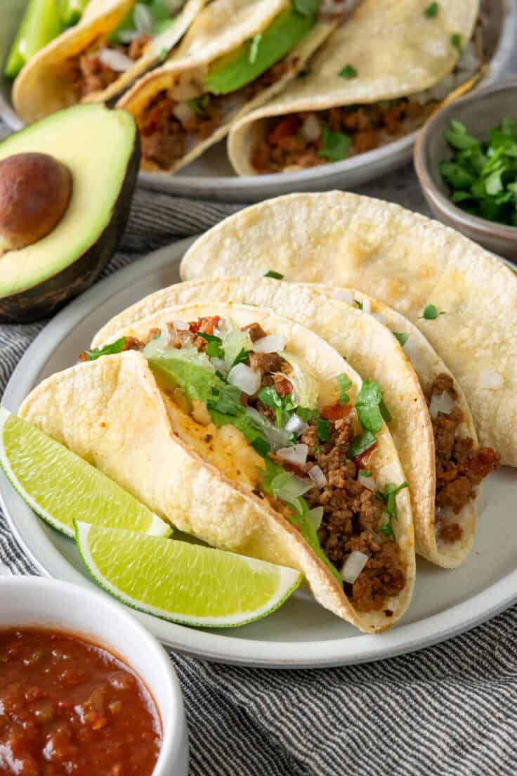 Plates of ground beef baked tacos with lime wedges served alongside bowls of cilantro, salsa, and avocado.
