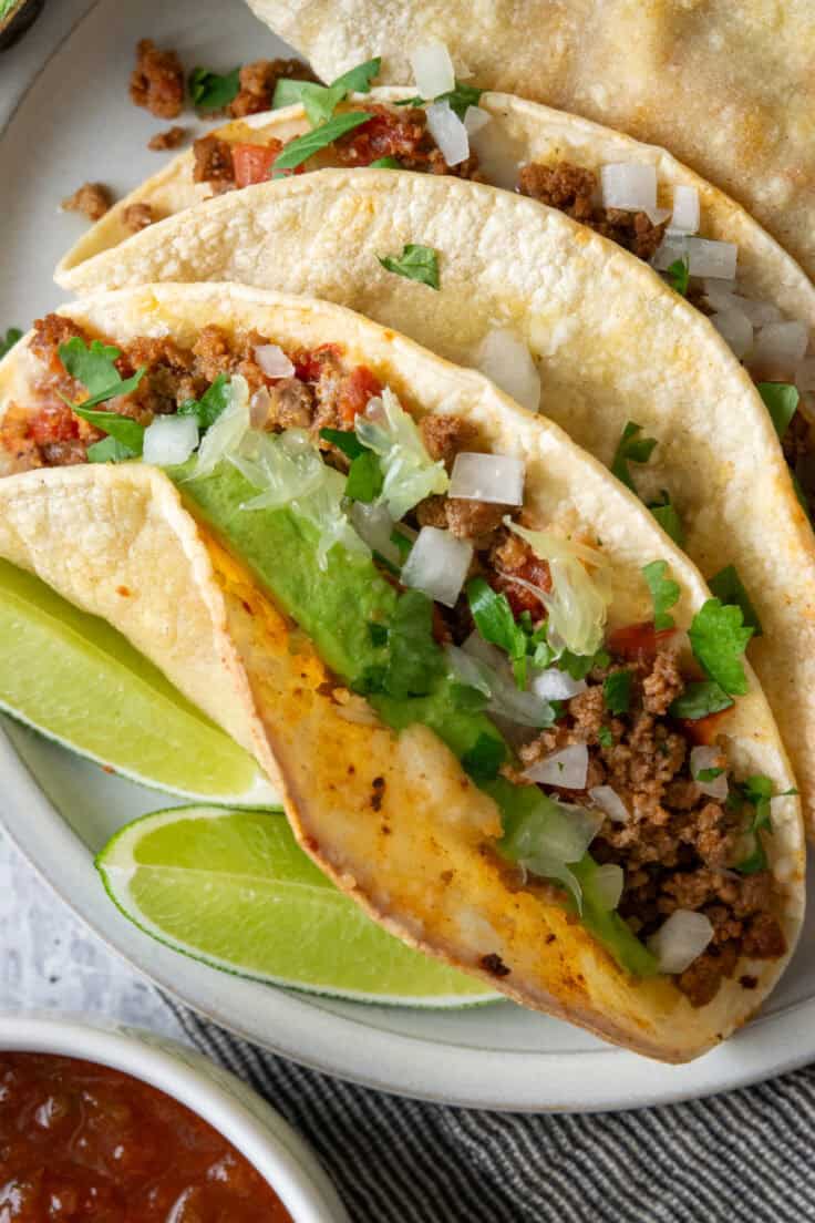 Close-up of a cheesy baked beef taco in a crispy corn tortilla with seasoned ground beef, onion, cilantro, avocado, and lime.