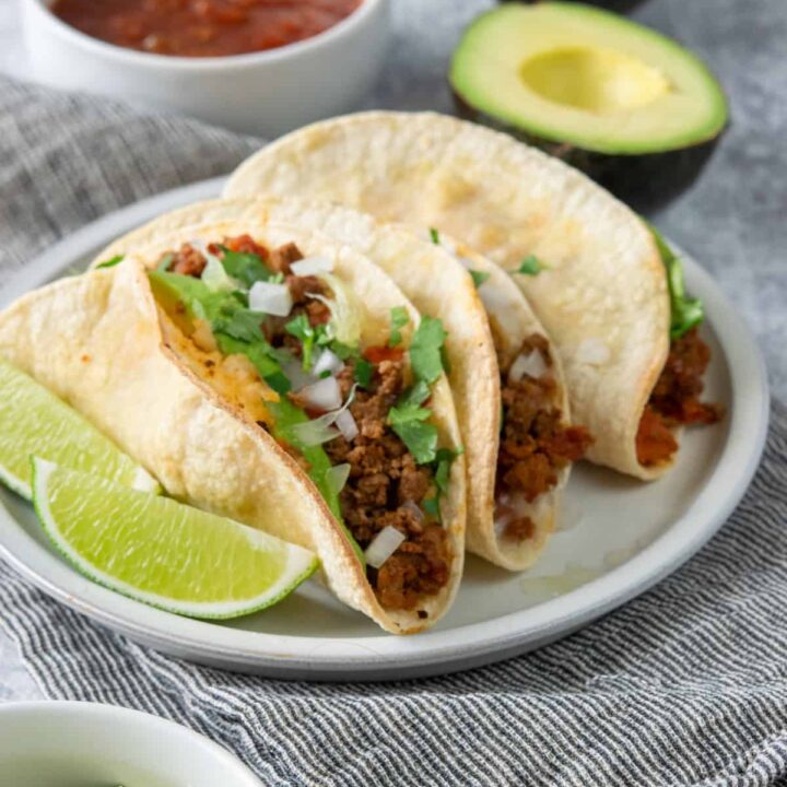 Crispy oven baked beef tacos topped with cheese, onion, cilantro, and avocado served with salsa, lime wedges, and avocado.
