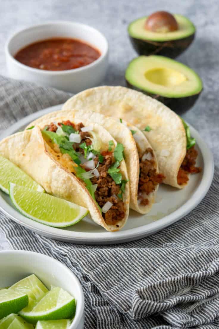 Crispy oven baked beef tacos topped with cheese, onion, cilantro, and avocado served with salsa, lime wedges, and avocado.