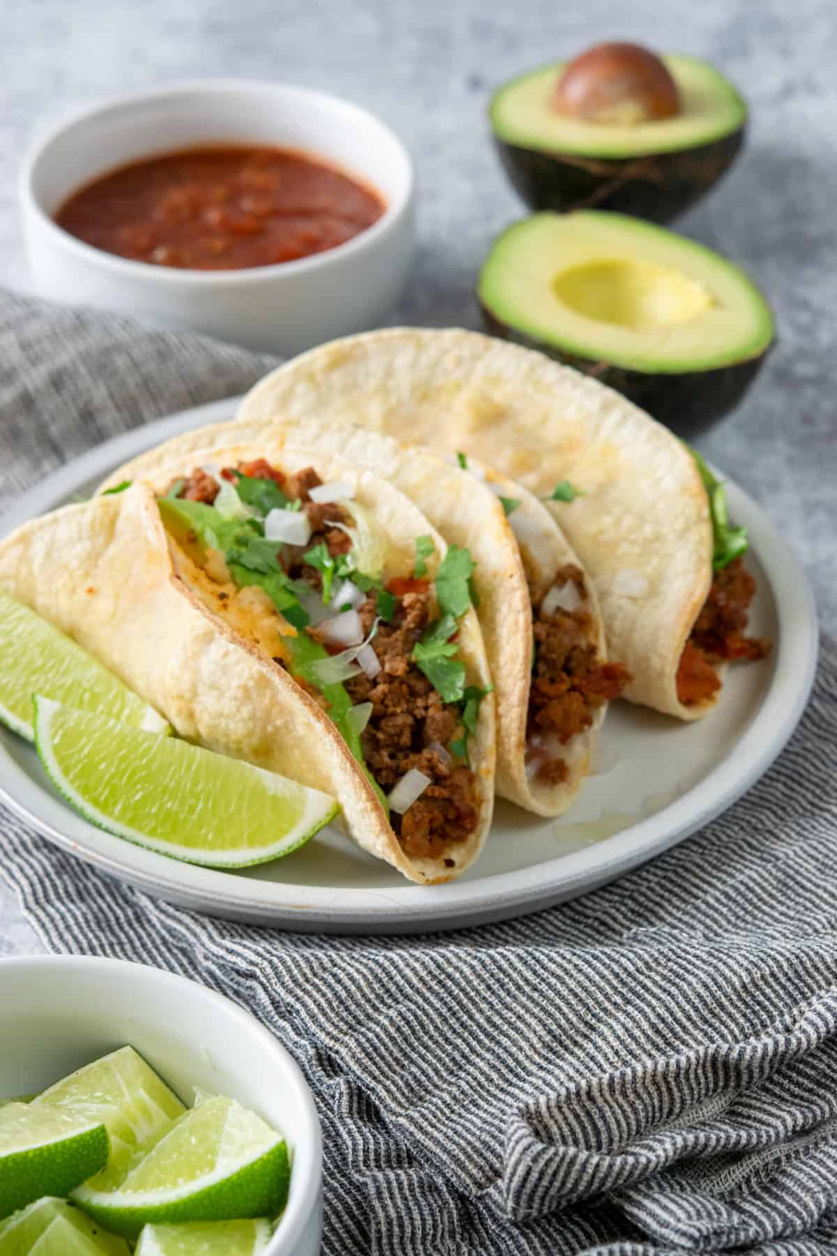 Crispy oven baked beef tacos topped with cheese, onion, cilantro, and avocado served with salsa, lime wedges, and avocado.