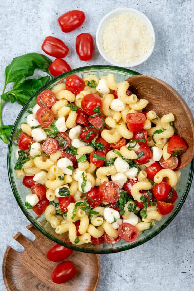 Caprese pasta salad in a green glass bowl with wooden serving spoons, styled for a fresh summer presentation.