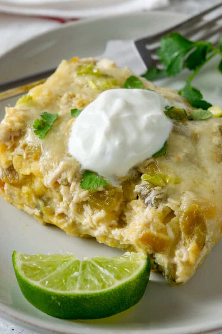 A hot, cheesy slice of chicken enchilada casserole on a plate with a lime garnish, topped with sour cream and cilantro.