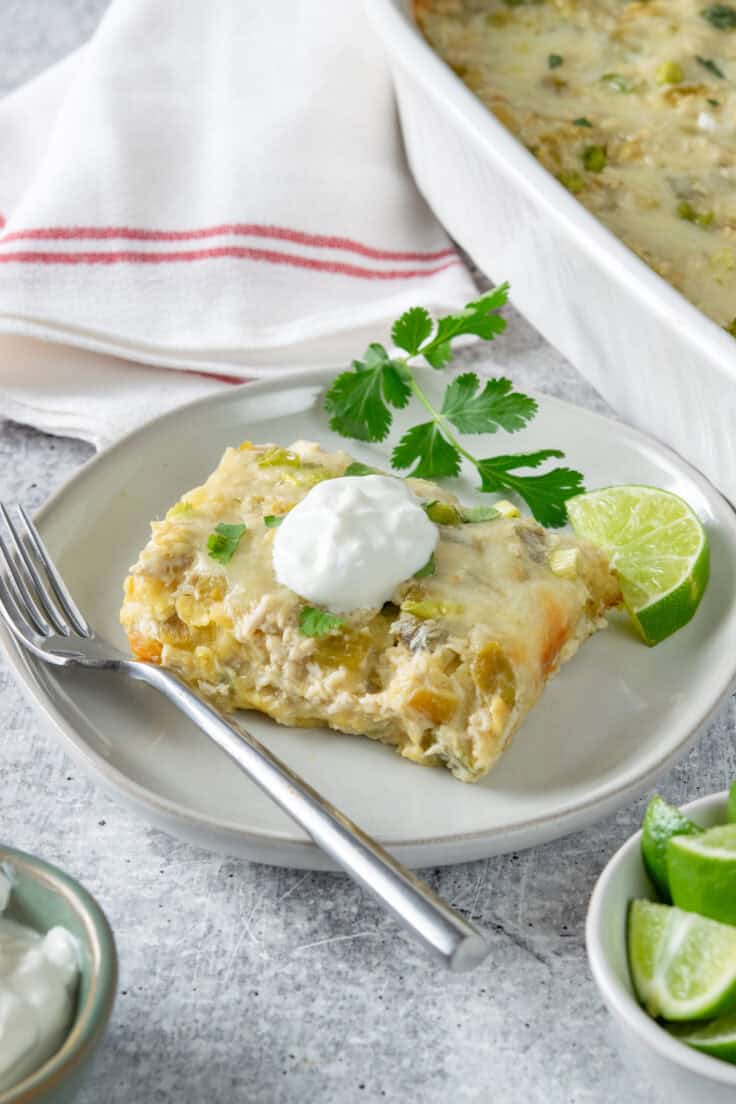Slice of green chile chicken enchilada casserole topped with sour cream and served with lime wedges and cilantro on plate.