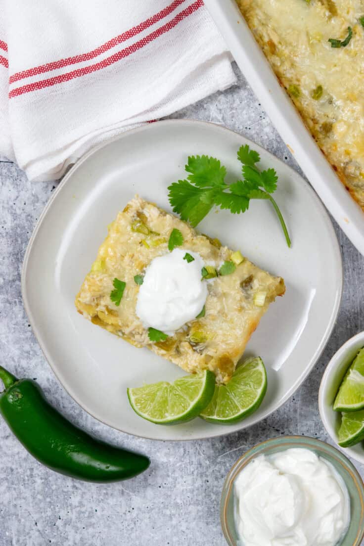 Overhead view of green chile chicken enchilada casserole slice with sour cream, jalapeño, lime wedges and casserole dish nearby.