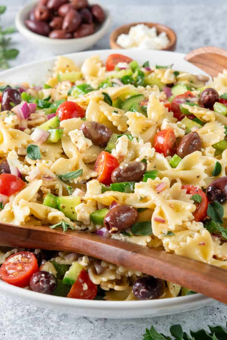 Serving bowl filled with Greek pasta salad featuring juicy tomatoes, feta cheese, olives, and vegetables with wooden serving utensils.
