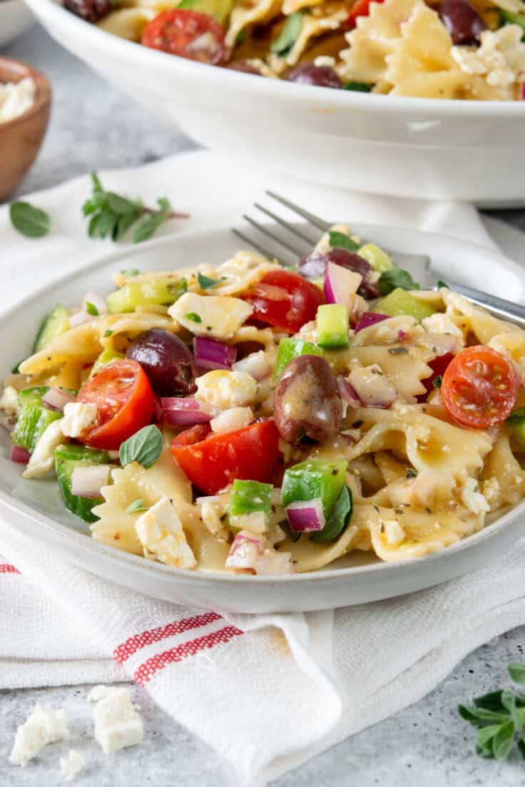 Plate of Greek pasta salad with vinaigrette dressing dripping from a Kalamata olive alongside pasta, feta, and vegetables.