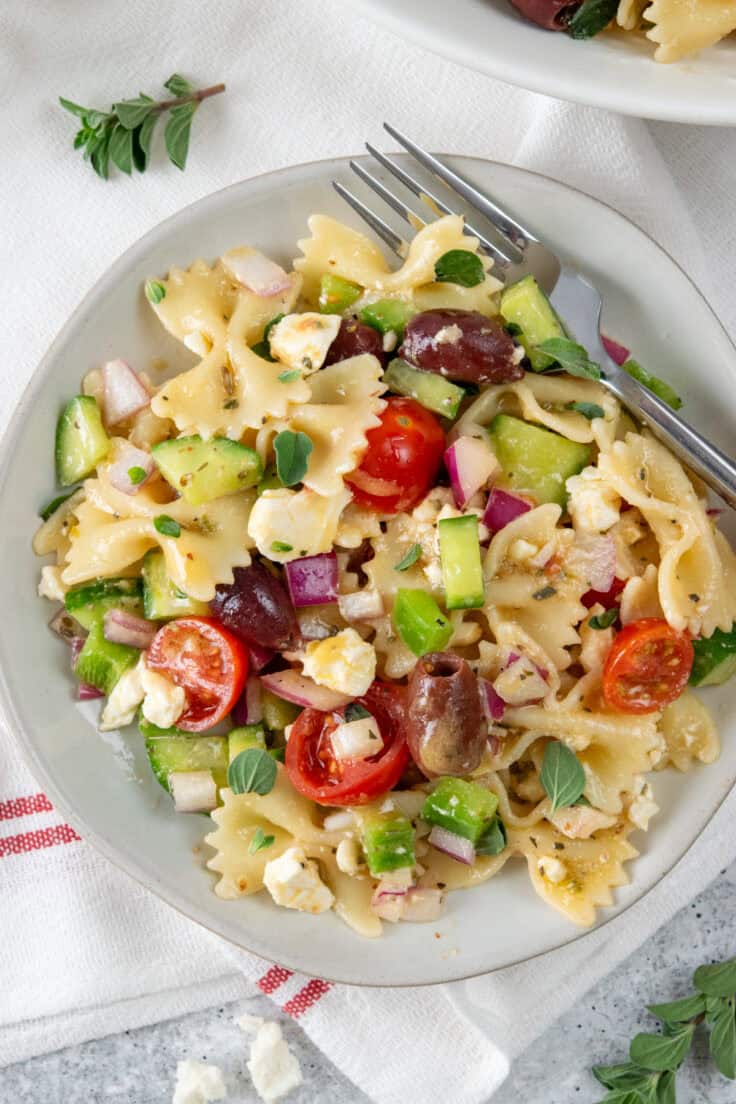 Overhead view of Greek pasta salad on a plate with fork showing bowtie pasta, cucumbers, tomatoes, olives, feta, and chopped vegetables in dressing.