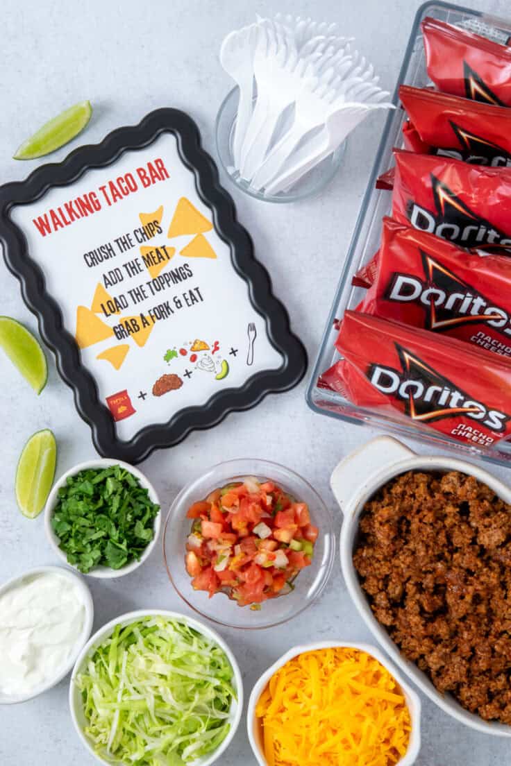 Overhead view of a make-your-own walking taco bar for a party, featuring a printable sign with instructions, a bucket of Doritos bags, plastic forks, seasoned taco meat, and bowls of taco toppings.