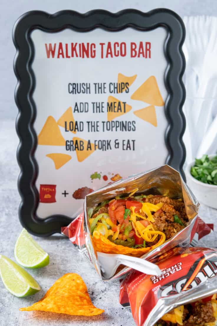 Walking taco chip bags laid on their sides to show seasoned meat and toppings inside, displayed next to a printable walking taco bar sign with simple step-by-step instructions for guests.