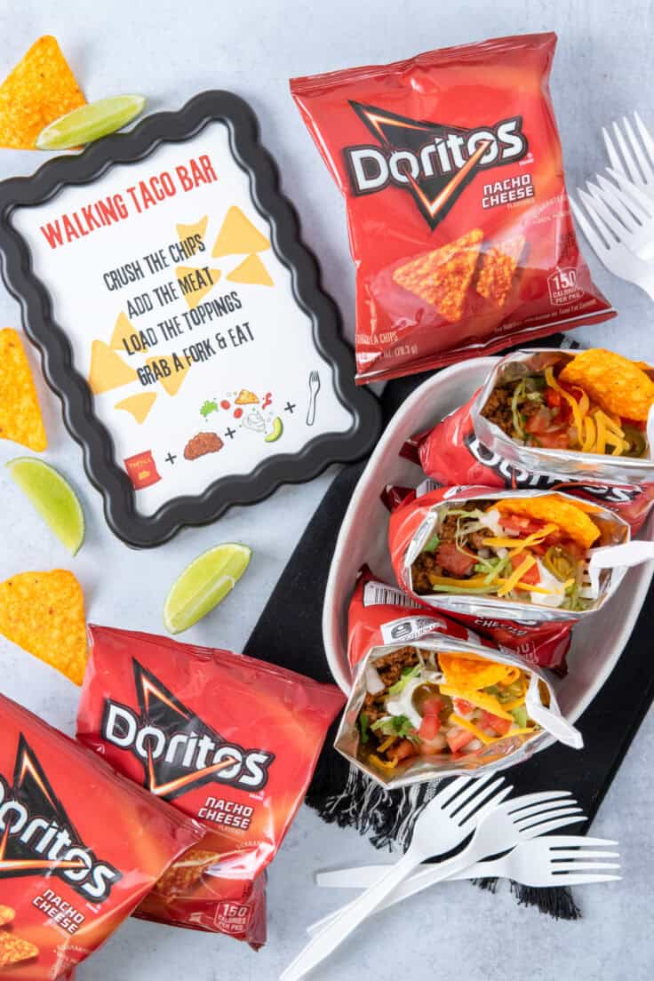 Overhead photo of a printable walking taco bar sign placed beside Doritos chip bags, plastic forks, and several prepared walking tacos with forks tucked inside the bags.