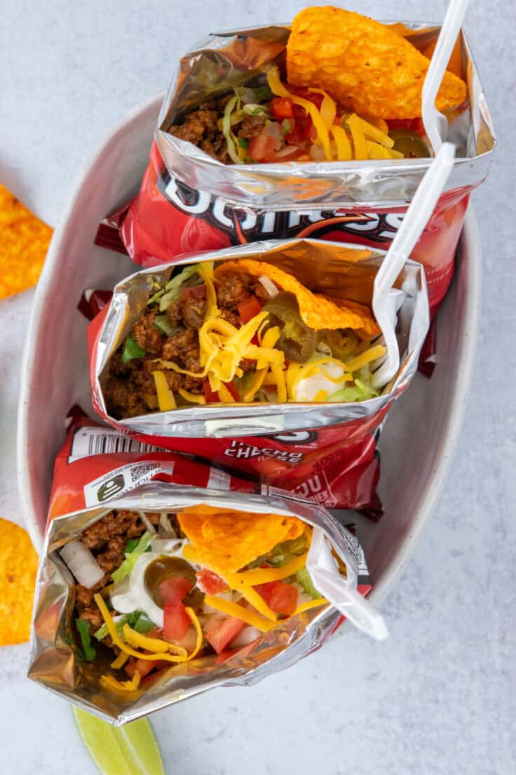 Multiple walking tacos assembled in Doritos bags, loaded with seasoned taco meat, shredded cheese, lettuce, tomatoes, sour cream, and a variety of colorful toppings.