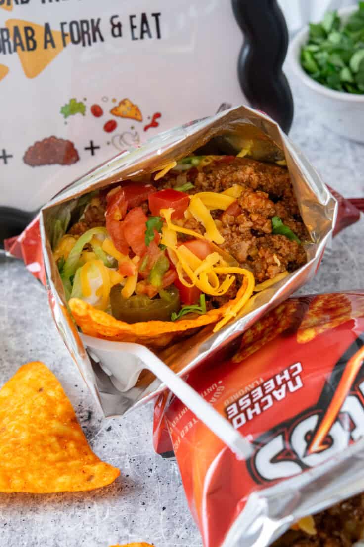 Walking taco in a Doritos chip bag laid on its side, showing layers of seasoned taco meat, crunchy chips, shredded cheese, sour cream, lettuce, onion, tomato, and sliced jalapeno.