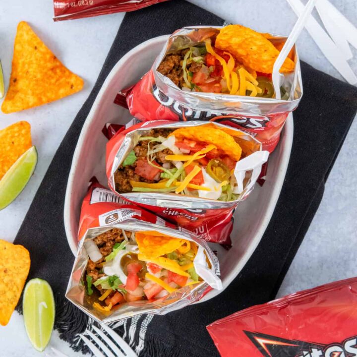 Overhead view of three walking tacos served in Doritos chip bags with forks, surrounded by extra chip bags, scattered chips, lime wedges, and plastic forks for a fun party setup.