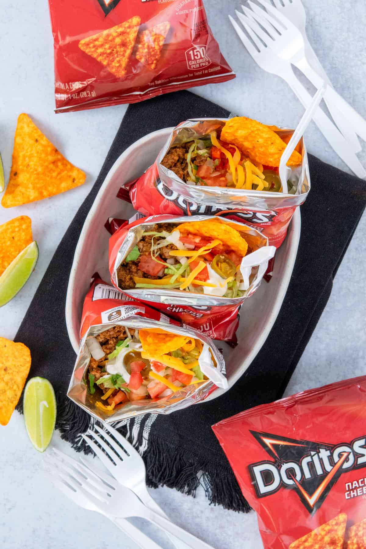 Overhead view of three walking tacos served in Doritos chip bags with forks, surrounded by extra chip bags, scattered chips, lime wedges, and plastic forks for a fun party setup.
