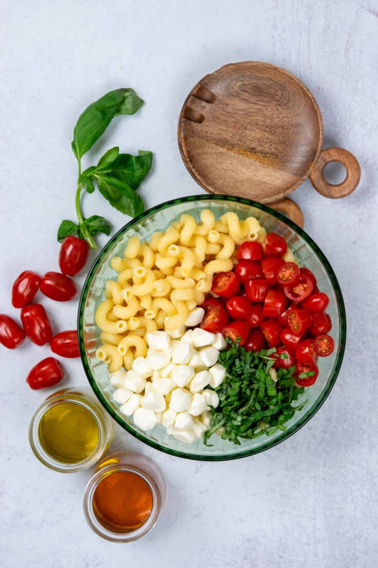 Bowl of caprese pasta salad before mixing with pasta, tomatoes, mozzarella, and basil alongside olive oil, vinegar, and wooden spoons.