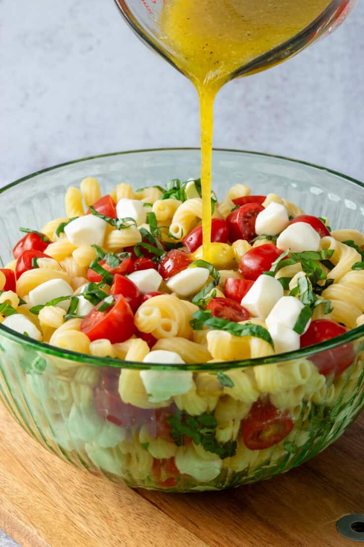 Large bowl of caprese pasta salad being tossed with tomatoes, mozzarella, basil, and dressing.