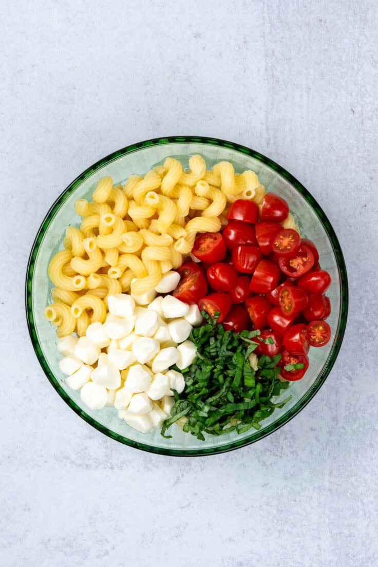 Cooked pasta, sliced tomatoes, basil, and pearl mozzarella balls in a large bowl ready to be tossed for caprese pasta salad.