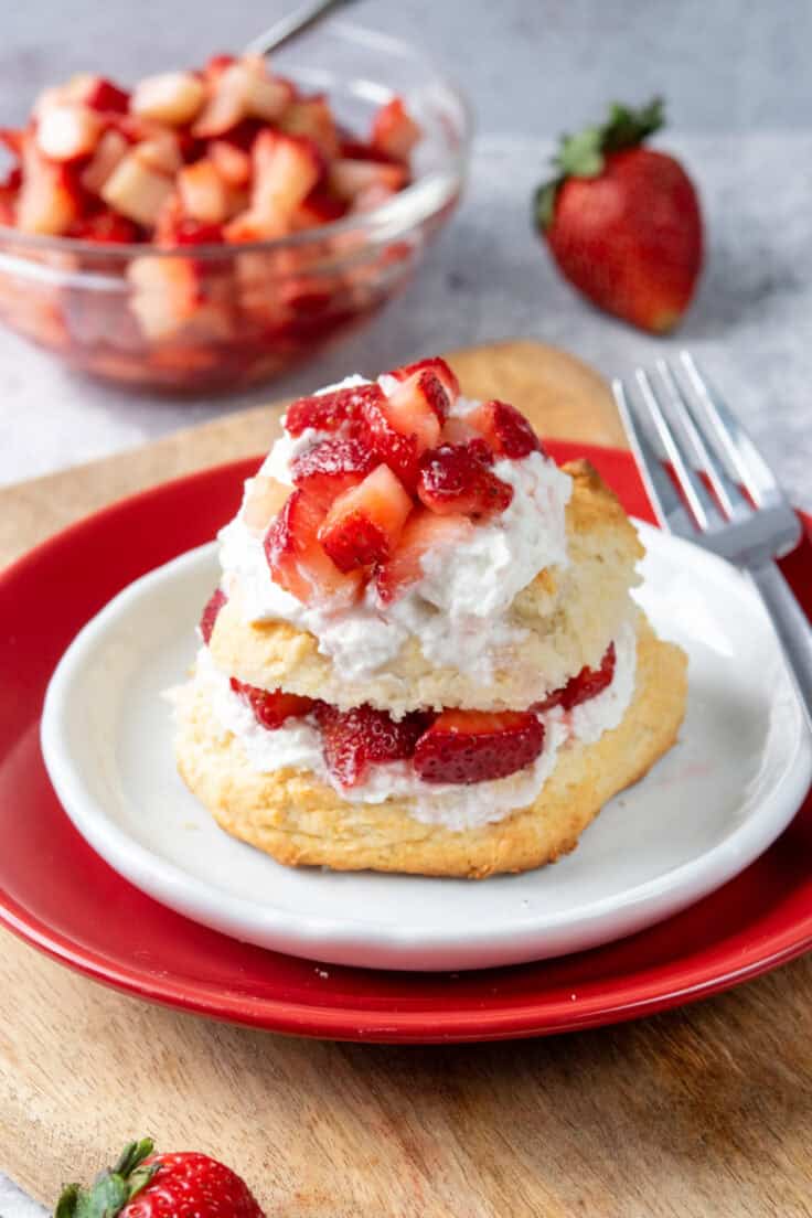 Bisquick strawberry shortcake layered with whipped cream, strawberries, and sweet strawberry syrup beside a bowl of macerated strawberries and a fresh strawberry.
