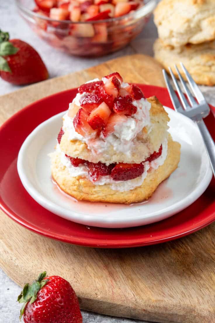 Bisquick strawberry shortcake piled high with whipped cream and juicy strawberries on a red and white dessert plate with a fork, surrounded by extra biscuits and a bowl of macerated strawberries in the background.