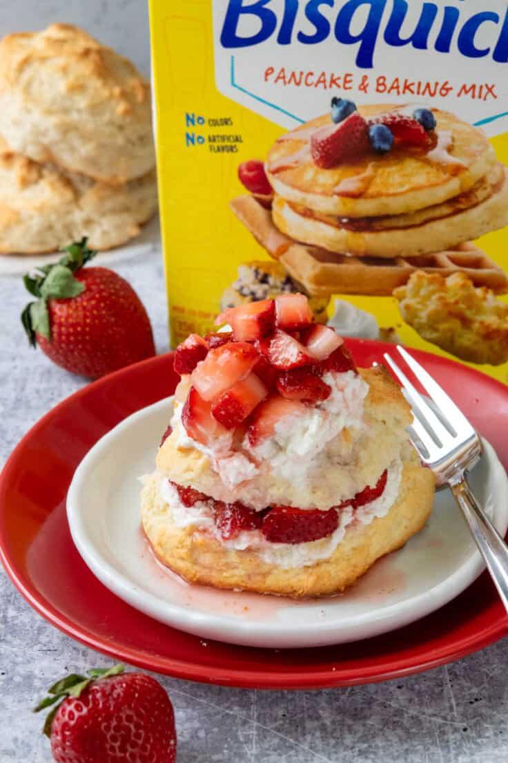 Bisquick strawberry shortcake stacked with whipped cream, juicy strawberries, and sweet strawberry syrup in front of a box of Bisquick baking mix and extra shortcake biscuits.