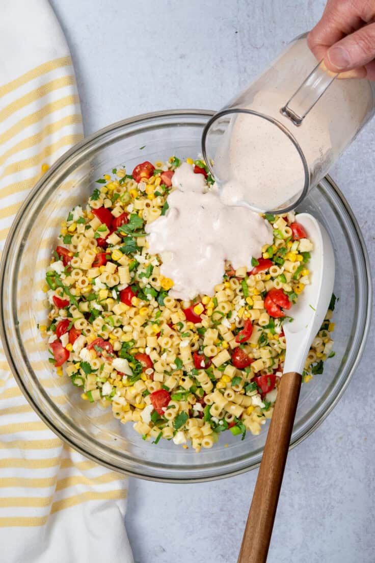 Creamy dressing being poured over mexican street corn pasta salad ingredients in a large bowl.