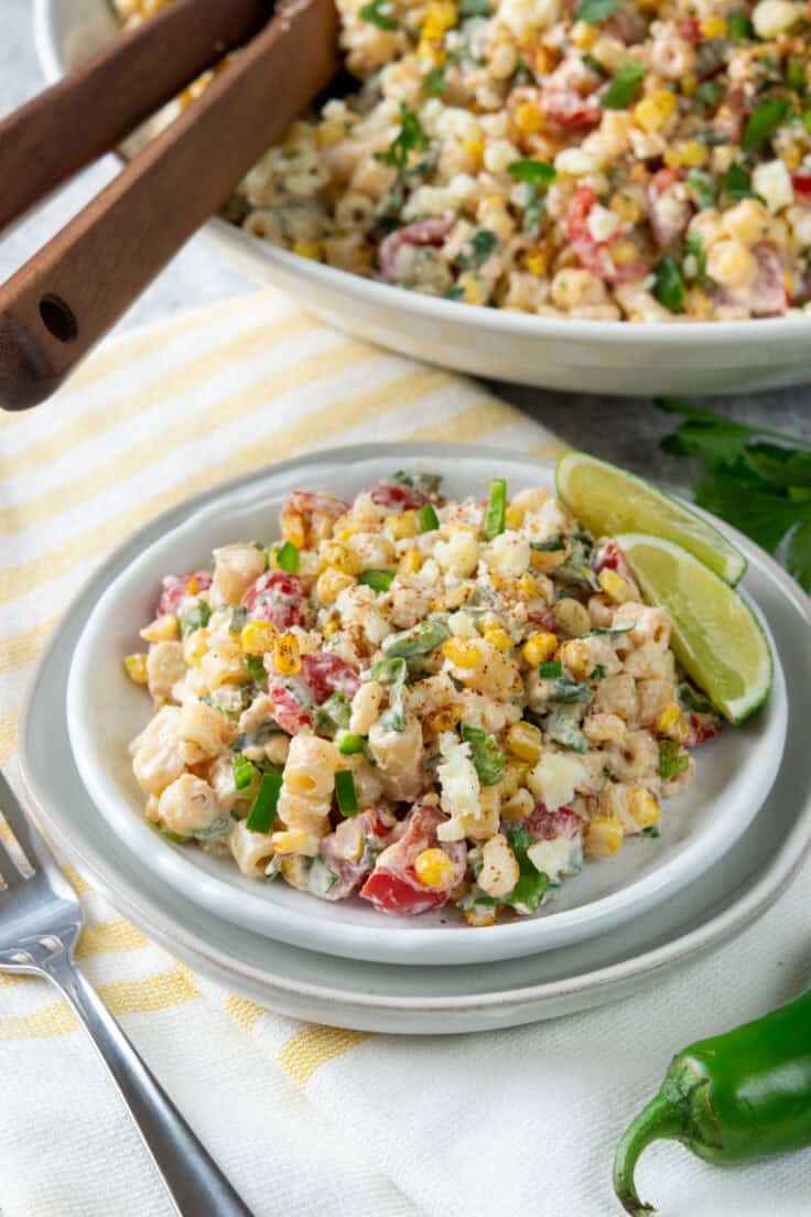 Mexican street corn pasta salad served on a small plate with a fork, alongside a large serving bowl with wooden spoons, lime wedge, and fresh jalapeño in the foreground.