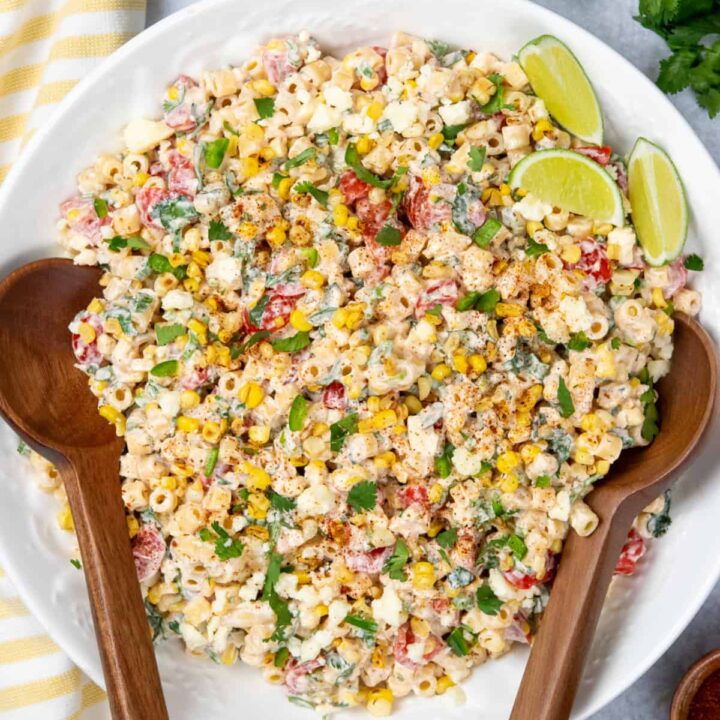 Large serving bowl of Mexican street corn pasta salad garnished with chili powder and surrounded by fresh cilantro, lime wedges, jalapeños, cotija cheese, and a bowl of chili powder.